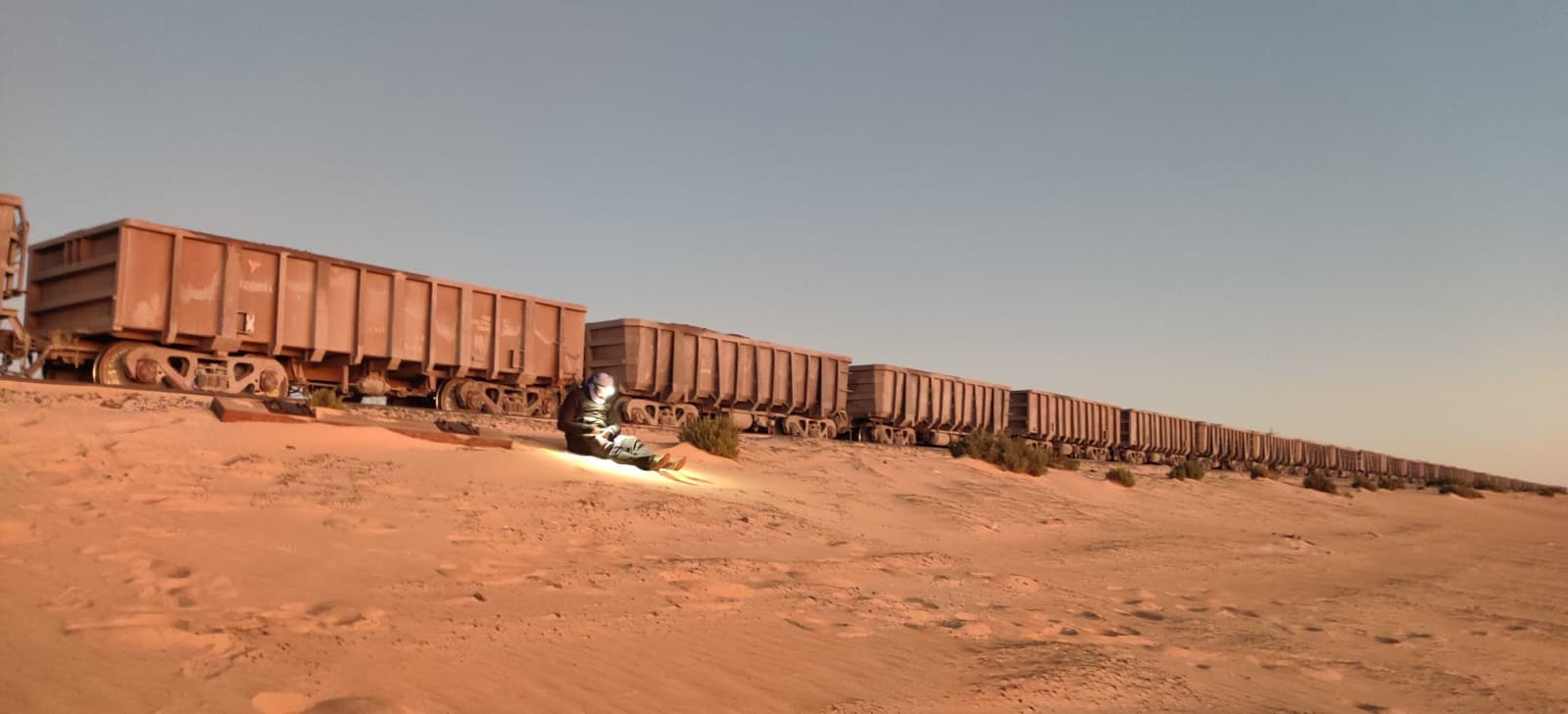 A traveller crouched beside the iron ore train wagons in the Mauritanian desert at dusk