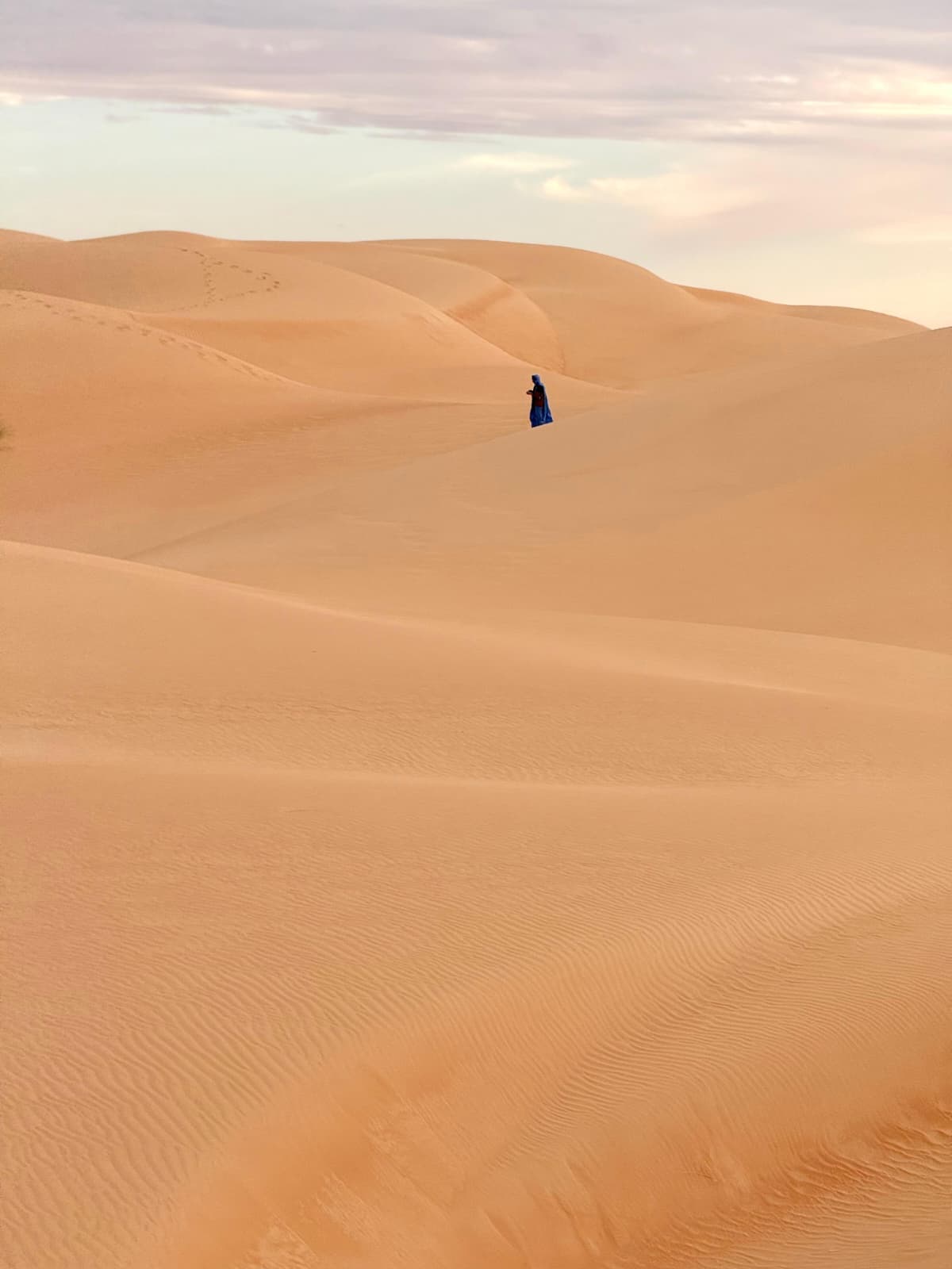 A lone figure in traditional dress walking across the Sahara dunes at dusk