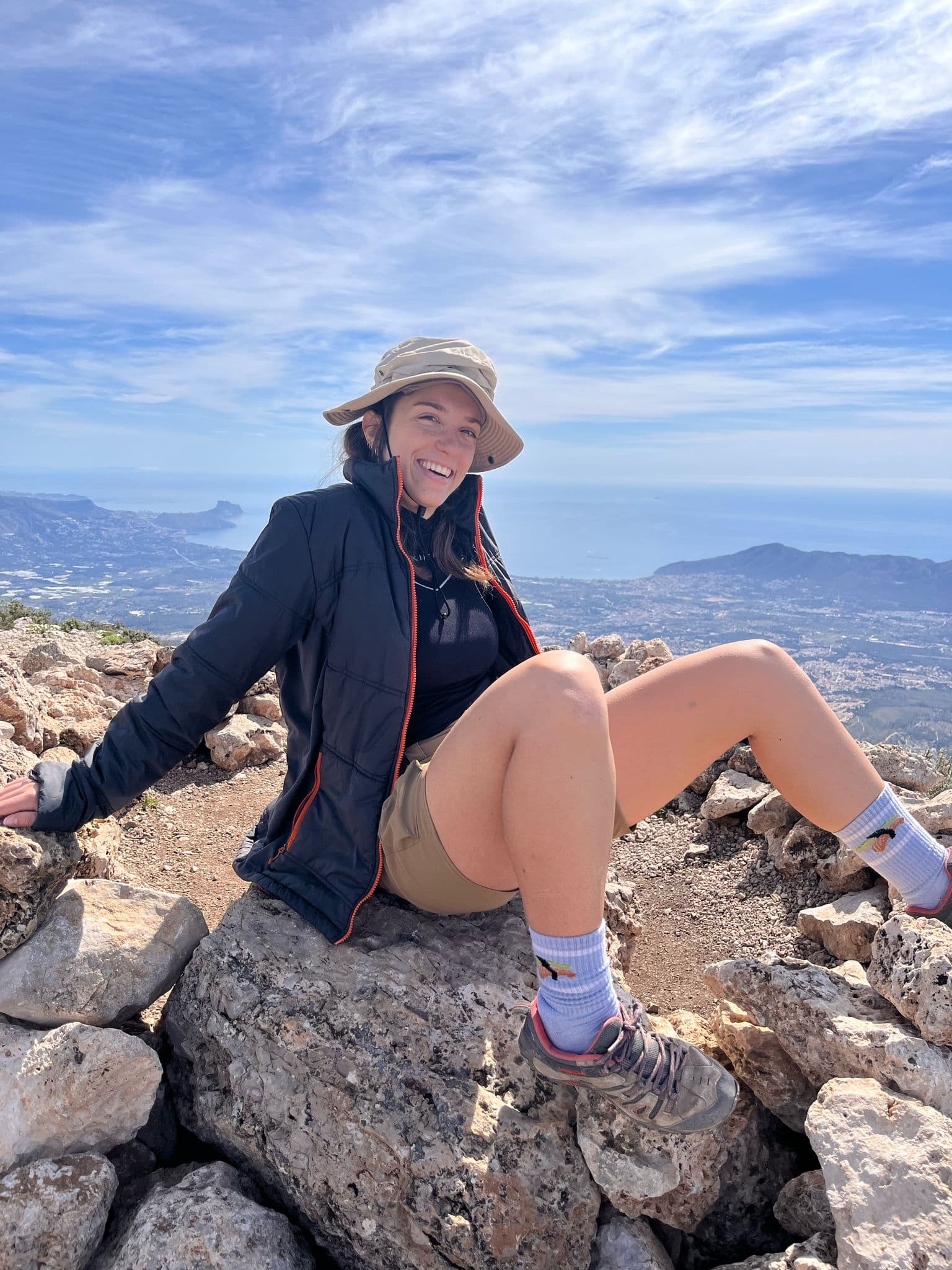 Sofia sitting on a rocky mountain summit above the coastline, mid-laugh, hiking gear on
